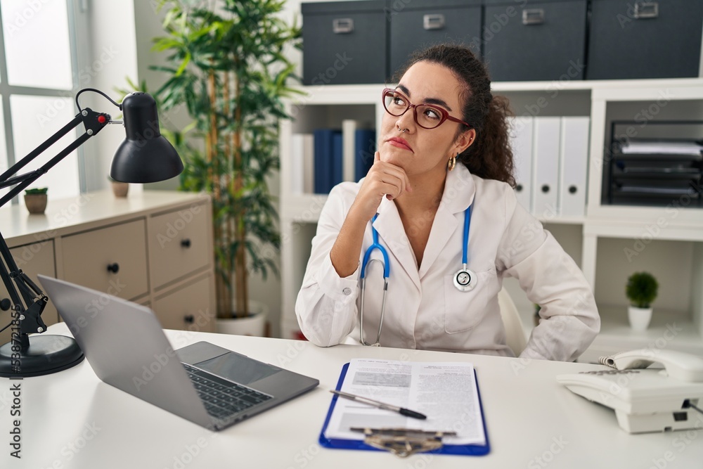 Young hispanic woman wearing doctor uniform and stethoscope with hand on chin thinking about question, pensive expression. smiling with thoughtful face. doubt concept. Young hispanic woman wearing doctor uniform and stethoscope with hand on chin thinking about question, pensive expression. smiling with thoughtful face. doubt concept.