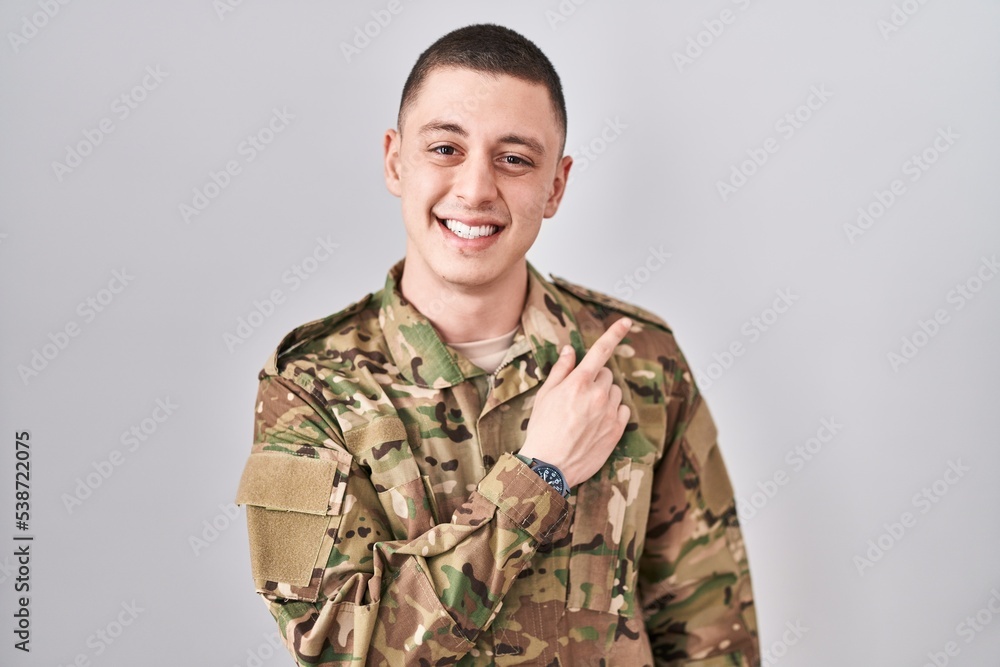 Young man wearing camouflage army uniform cheerful with a smile of face pointing with hand and finger up to the side with happy and natural expression on face