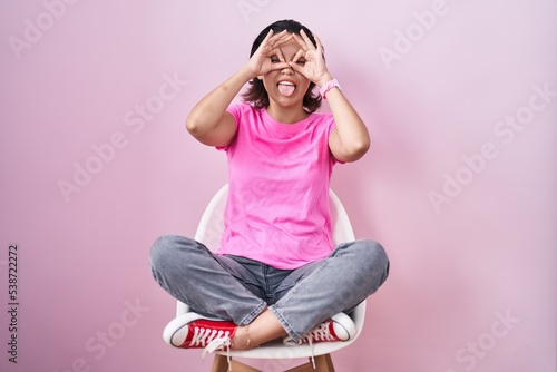 Hispanic young woman sitting on chair over pink background doing ok gesture like binoculars sticking tongue out, eyes looking through fingers. crazy expression. © Krakenimages.com