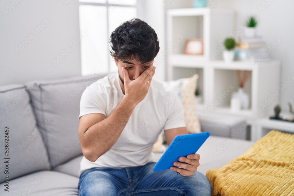 Hispanic man with beard using touchpad sitting on the sofa shocked covering mouth with hands for mistake. secret concept. Hispanic man with beard using touchpad sitting on the sofa shocked covering mouth with hands for mistake. secret concept.
