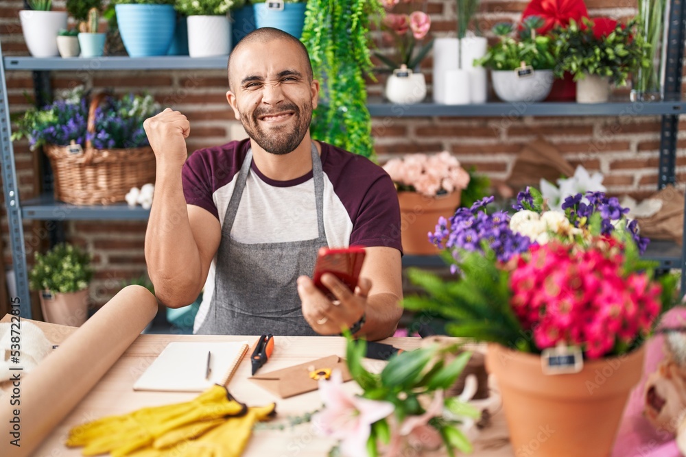 Hispanic man with beard working at florist shop with smartphone screaming proud, celebrating victory and success very excited with raised arm Hispanic man with beard working at florist shop with smartphone screaming proud, celebrating victory and success very excited with raised arm