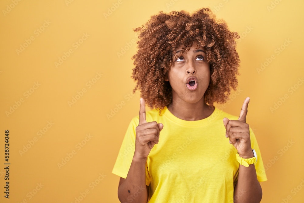 Young hispanic woman with curly hair standing over yellow background amazed and surprised looking up and pointing with fingers and raised arms. Young hispanic woman with curly hair standing over yellow background amazed and surprised looking up and pointing with fingers and raised arms.