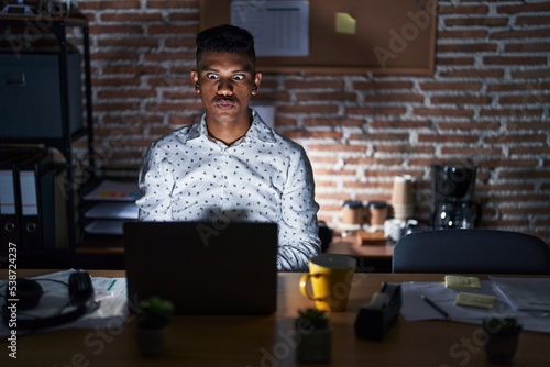 Young hispanic man working at the office at night making fish face with lips, crazy and comical gesture. funny expression.