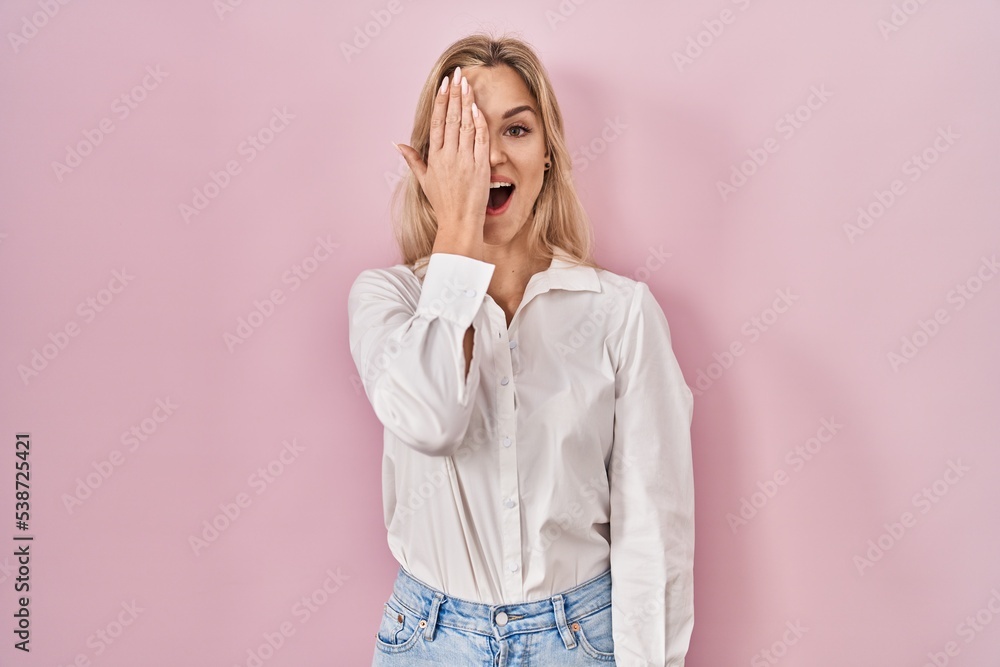 Young caucasian woman wearing casual white shirt over pink background covering one eye with hand, confident smile on face and surprise emotion. Young caucasian woman wearing casual white shirt over pink background covering one eye with hand, confident smile on face and surprise emotion.