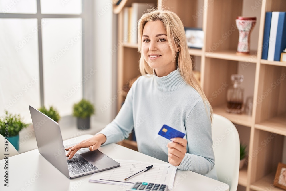 Fototapeta premium Young blonde woman using laptop and credit card sitting on table at home
