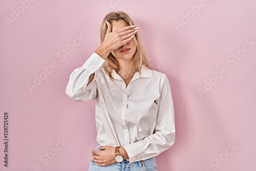 Young caucasian woman wearing casual white shirt over pink background covering eyes with hand, looking serious and sad. sightless, hiding and rejection concept © Krakenimages.com