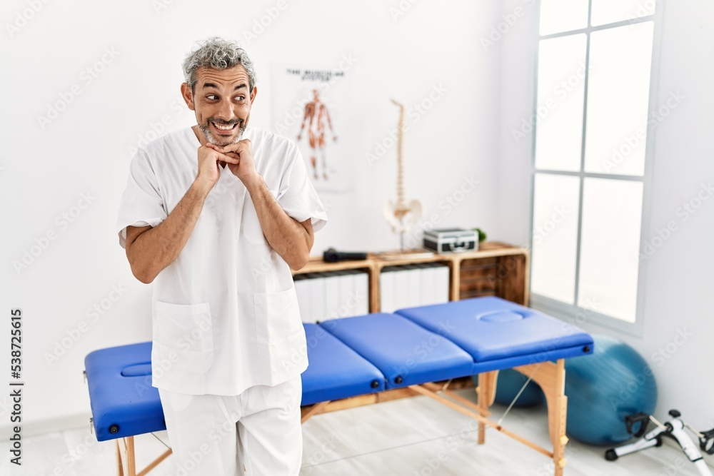 Middle age hispanic therapist man working at pain recovery clinic laughing nervous and excited with hands on chin looking to the side Middle age hispanic therapist man working at pain recovery clinic laughing nervous and excited with hands on chin looking to the side