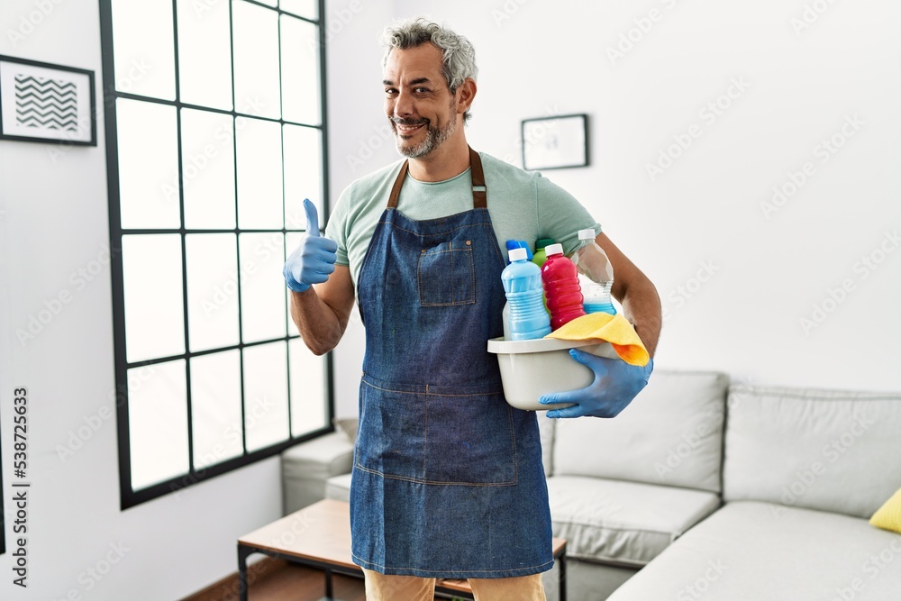 Middle age hispanic man wearing cleaner apron holding cleaning products smiling happy and positive, thumb up doing excellent and approval sign