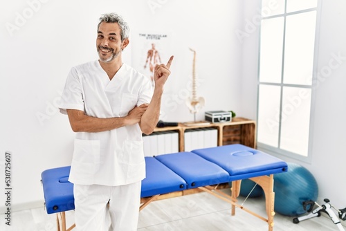 Middle age hispanic therapist man working at pain recovery clinic with a big smile on face  pointing with hand finger to the side looking at the camera.
