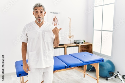 Middle age hispanic therapist man working at pain recovery clinic surprised pointing with hand finger to the side  open mouth amazed expression.