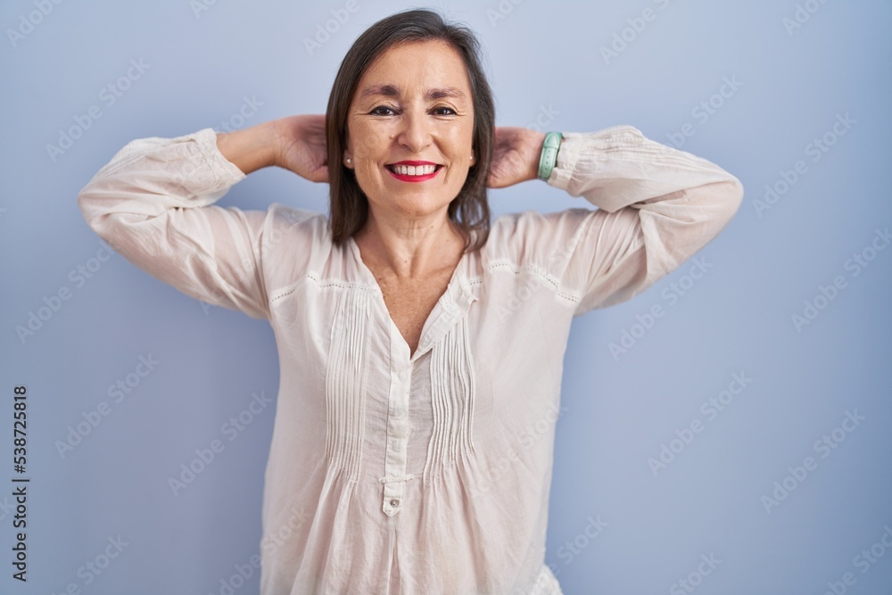 Middle age hispanic woman standing over blue background relaxing and stretching, arms and hands behind head and neck smiling happy Middle age hispanic woman standing over blue background relaxing and stretching, arms and hands behind head and neck smiling happy
