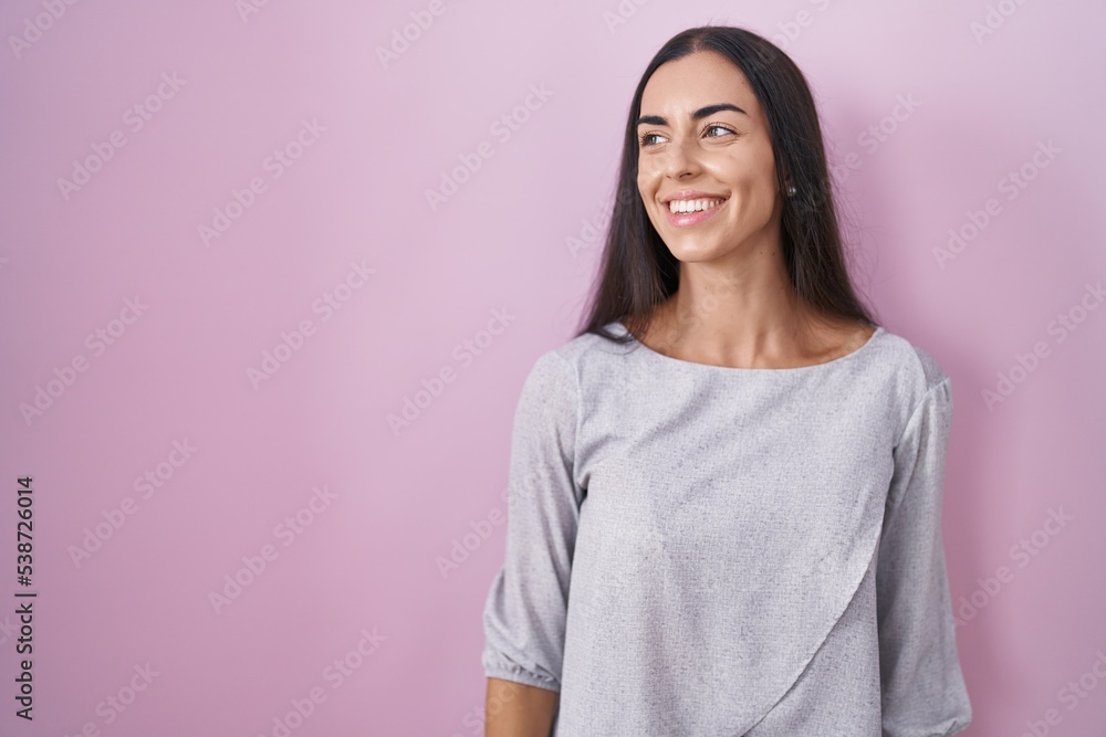 Young brunette woman standing over pink background looking away to side with smile on face, natural expression. laughing confident. Young brunette woman standing over pink background looking away to side with smile on face, natural expression. laughing confident.