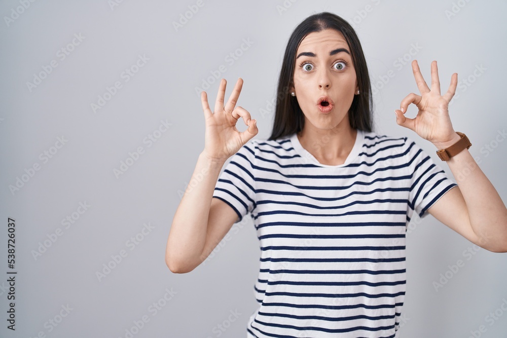 Young brunette woman wearing striped t shirt looking surprised and shocked doing ok approval symbol with fingers. crazy expression Young brunette woman wearing striped t shirt looking surprised and shocked doing ok approval symbol with fingers. crazy expression