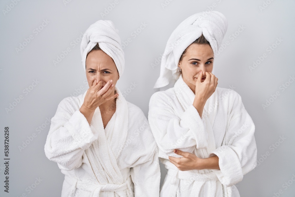 Middle age woman and daughter wearing white bathrobe and towel smelling something stinky and disgusting, intolerable smell, holding breath with fingers on nose. bad smell Middle age woman and daughter wearing white bathrobe and towel smelling something stinky and disgusting, intolerable smell, holding breath with fingers on nose. bad smell