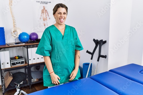 Young hispanic woman wearing physiotherapist uniform standing at clinic sticking tongue out happy with funny expression. emotion concept.