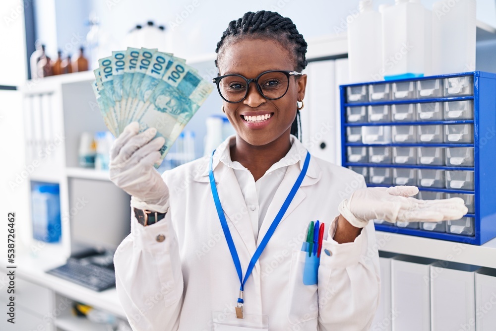 African woman with braids working at scientist laboratory holding money celebrating achievement with happy smile and winner expression with raised hand
