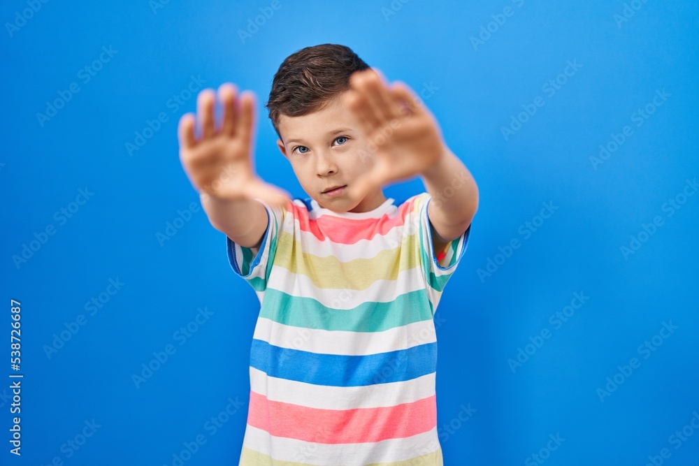 Young caucasian kid standing over blue background doing frame using hands palms and fingers, camera perspective Young caucasian kid standing over blue background doing frame using hands palms and fingers, camera perspective