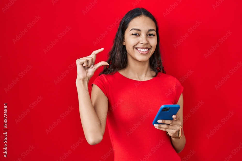 Naklejka premium Young brazilian woman using smartphone over red background smiling and confident gesturing with hand doing small size sign with fingers looking and the camera. measure concept.
