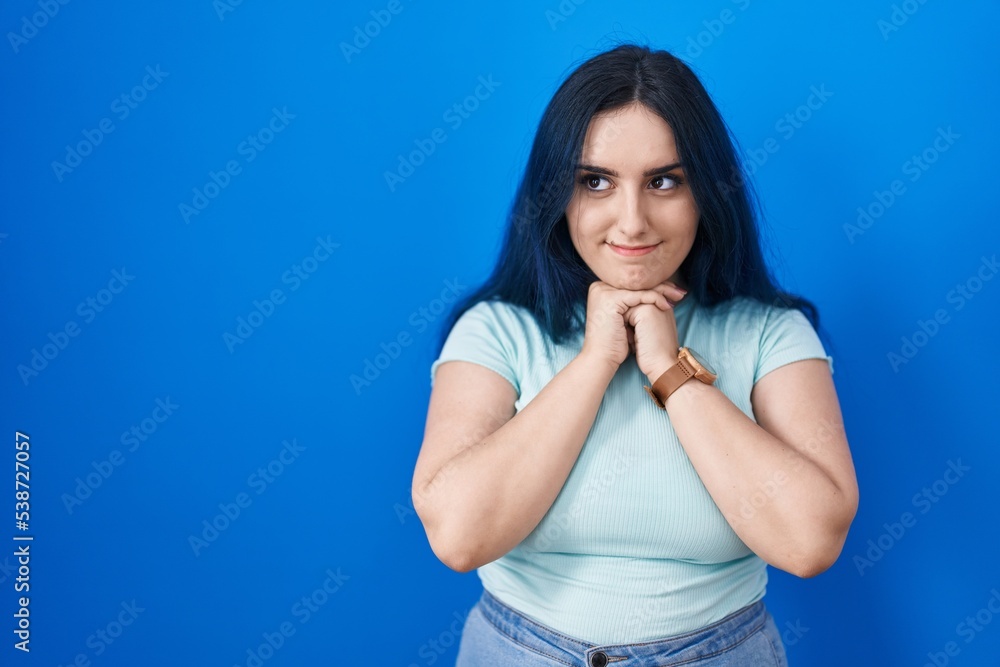 Young modern girl with blue hair standing over blue background laughing nervous and excited with hands on chin looking to the side Young modern girl with blue hair standing over blue background laughing nervous and excited with hands on chin looking to the side