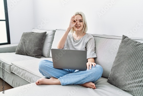 Young caucasian woman using laptop at home sitting on the sofa doing ok gesture with hand smiling, eye looking through fingers with happy face.