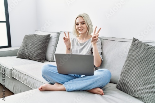 Young caucasian woman using laptop at home sitting on the sofa smiling looking to the camera showing fingers doing victory sign. number two.