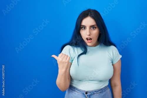 Young modern girl with blue hair standing over blue background surprised pointing with hand finger to the side, open mouth amazed expression.