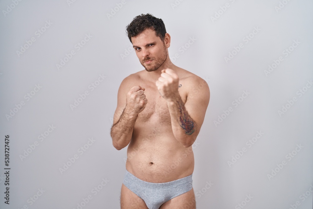 Young hispanic man standing shirtless wearing underware ready to fight with fist defense gesture, angry and upset face, afraid of problem