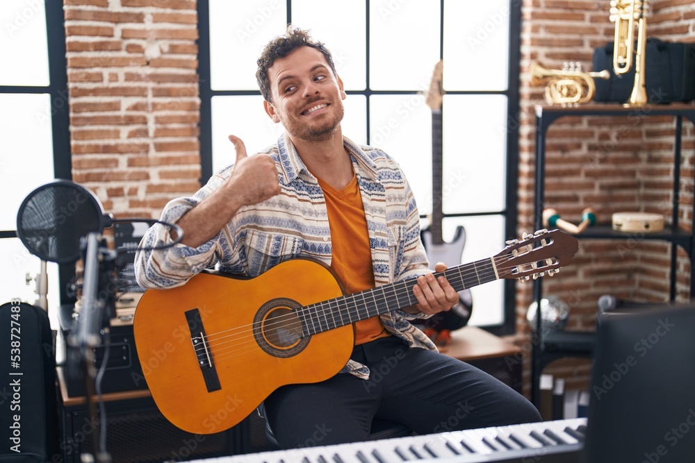 Fototapeta premium Young hispanic man playing classic guitar at music studio pointing thumb up to the side smiling happy with open mouth