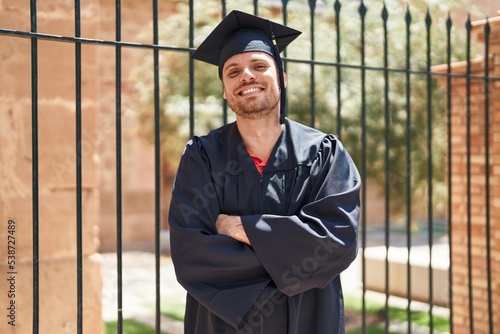 Young hispanic man wearing graduate uniform standing with arms crossed gesture at street