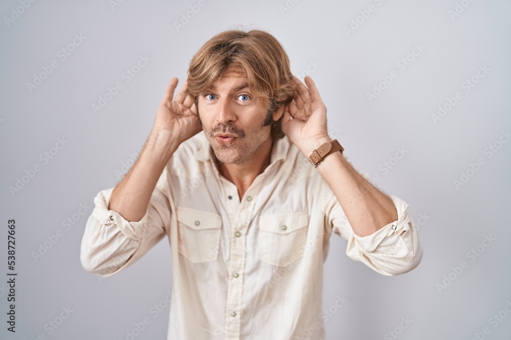 Middle age man standing over isolated background trying to hear both hands on ear gesture, curious for gossip. hearing problem, deaf Middle age man standing over isolated background trying to hear both hands on ear gesture, curious for gossip. hearing problem, deaf