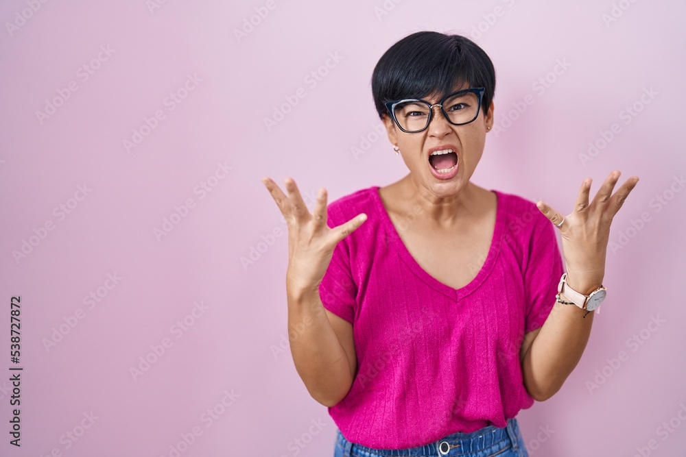 Young asian woman with short hair standing over pink background crazy and mad shouting and yelling with aggressive expression and arms raised. frustration concept. Young asian woman with short hair standing over pink background crazy and mad shouting and yelling with aggressive expression and arms raised. frustration concept.