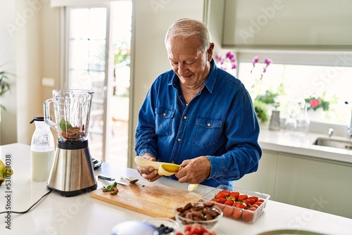 Senior man smiling confident holding peeled banana at kitchen