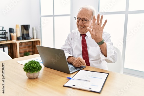 Senior man working at the office using computer laptop waiving saying hello happy and smiling, friendly welcome gesture