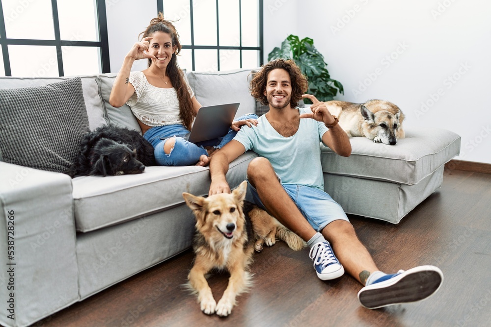 Young hispanic couple with dogs relaxing at home smiling and confident gesturing with hand doing small size sign with fingers looking and the camera. measure concept.