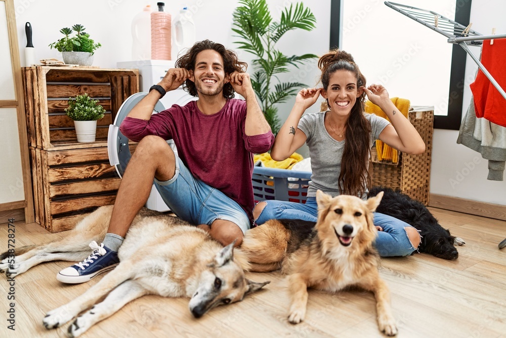 Young hispanic couple doing laundry with dogs smiling pulling ears with fingers, funny gesture. audition problem
