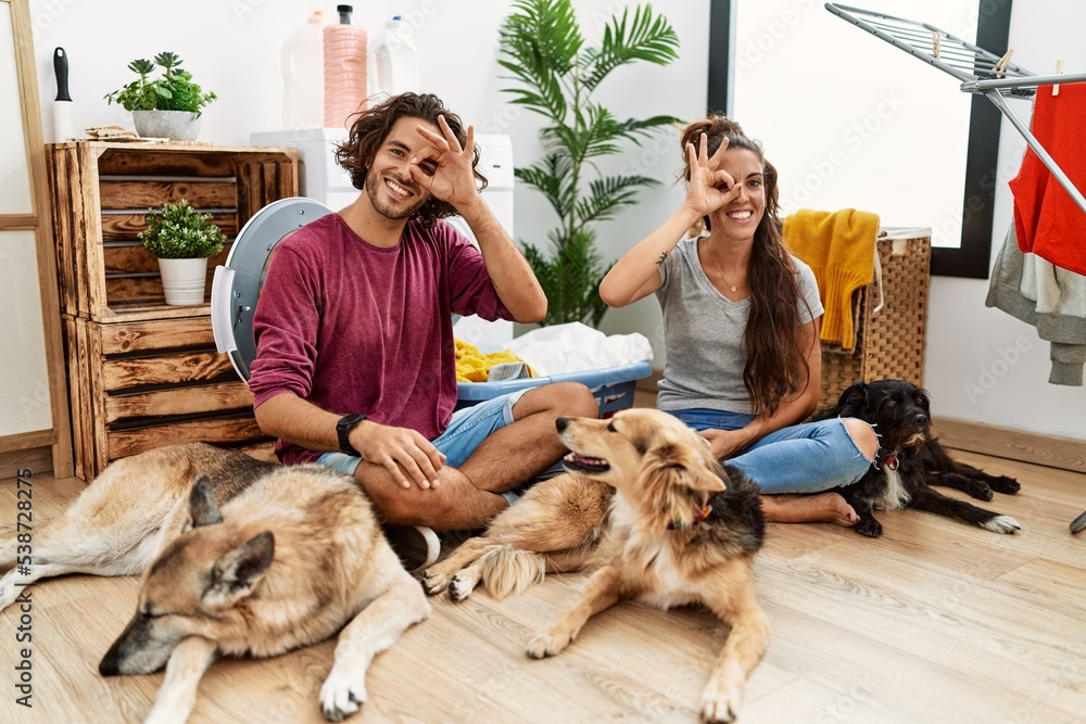 Obraz premium Young hispanic couple doing laundry with dogs doing ok gesture with hand smiling, eye looking through fingers with happy face.