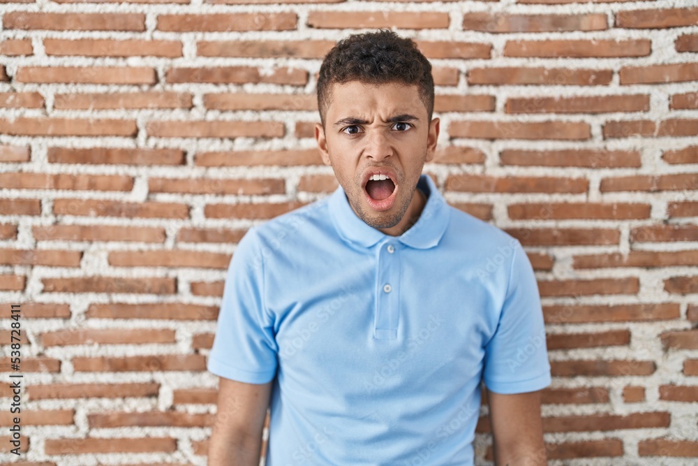 Brazilian young man standing over brick wall in shock face, looking skeptical and sarcastic, surprised with open mouth Brazilian young man standing over brick wall in shock face, looking skeptical and sarcastic, surprised with open mouth