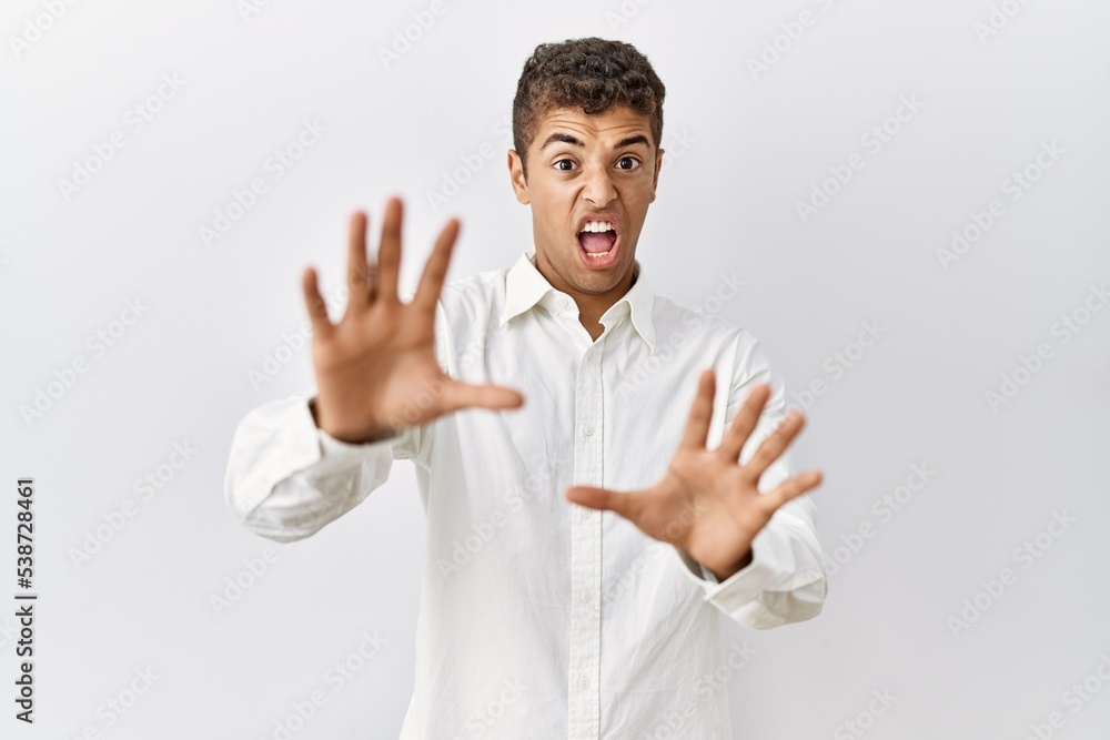 Young handsome hispanic man standing over isolated background afraid and terrified with fear expression stop gesture with hands, shouting in shock. panic concept. Young handsome hispanic man standing over isolated background afraid and terrified with fear expression stop gesture with hands, shouting in shock. panic concept.