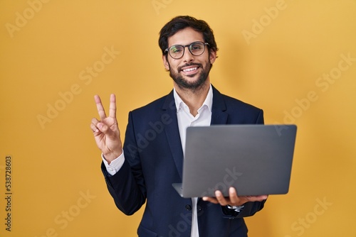 Handsome latin man working using computer laptop smiling with happy face winking at the camera doing victory sign with fingers. number two.