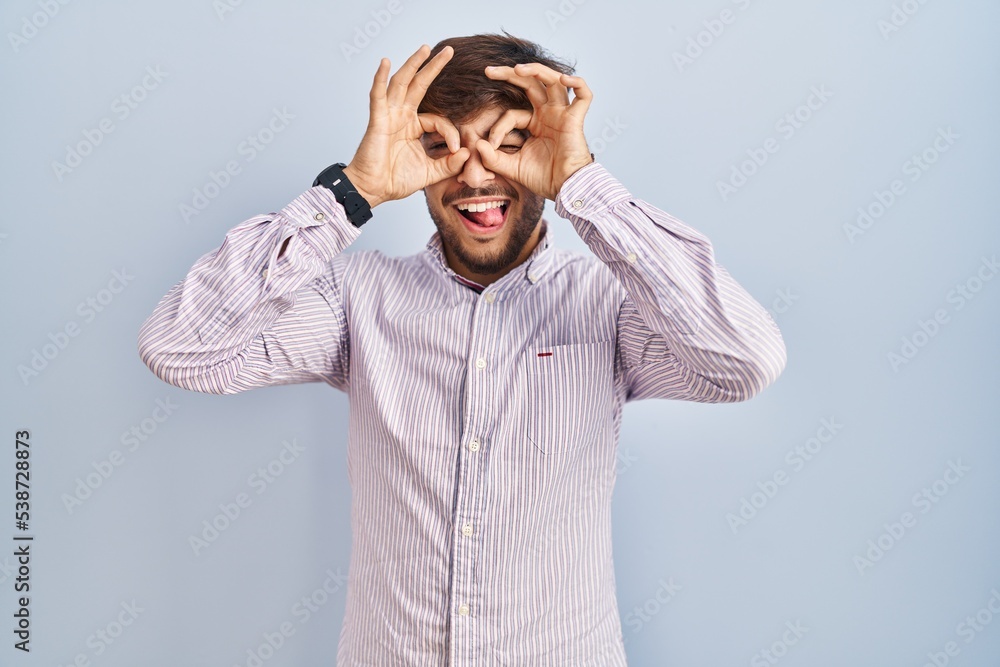 Arab man with beard standing over blue background doing ok gesture like binoculars sticking tongue out, eyes looking through fingers. crazy expression. Arab man with beard standing over blue background doing ok gesture like binoculars sticking tongue out, eyes looking through fingers. crazy expression.