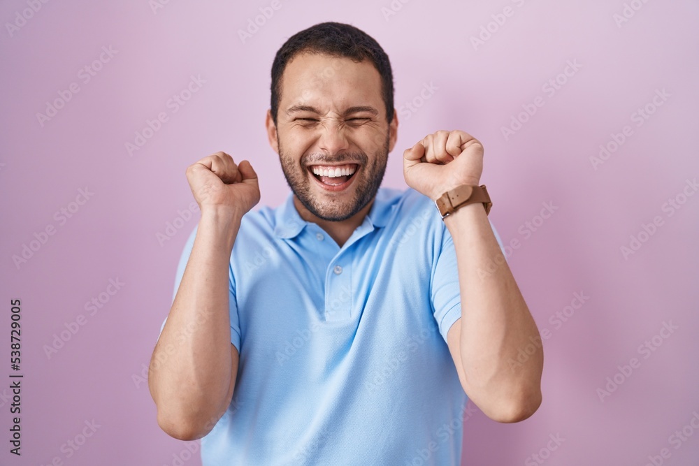 Hispanic man standing over pink background excited for success with arms raised and eyes closed celebrating victory smiling. winner concept. Hispanic man standing over pink background excited for success with arms raised and eyes closed celebrating victory smiling. winner concept.