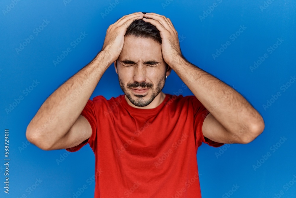 Young hispanic man with beard wearing red t shirt over blue background suffering from headache desperate and stressed because pain and migraine. hands on head. Young hispanic man with beard wearing red t shirt over blue background suffering from headache desperate and stressed because pain and migraine. hands on head.