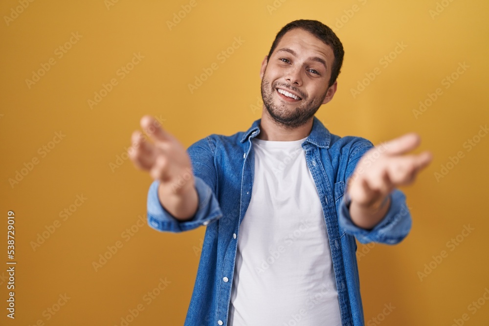 Hispanic man standing over yellow background smiling cheerful offering hands giving assistance and acceptance. Hispanic man standing over yellow background smiling cheerful offering hands giving assistance and acceptance.