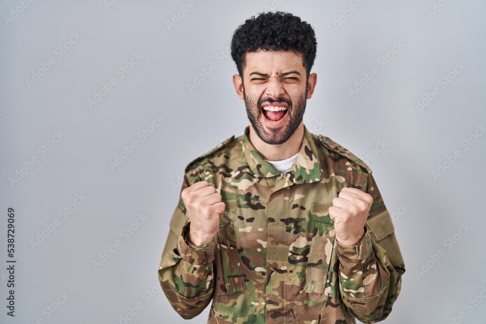 Arab man wearing camouflage army uniform excited for success with arms raised and eyes closed celebrating victory smiling. winner concept.