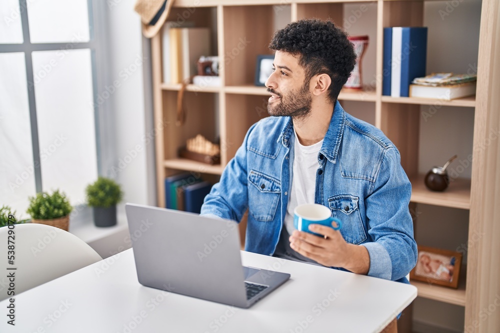 Young arab man using laptop drinking coffe sitting on table at home Young arab man using laptop drinking coffe sitting on table at home