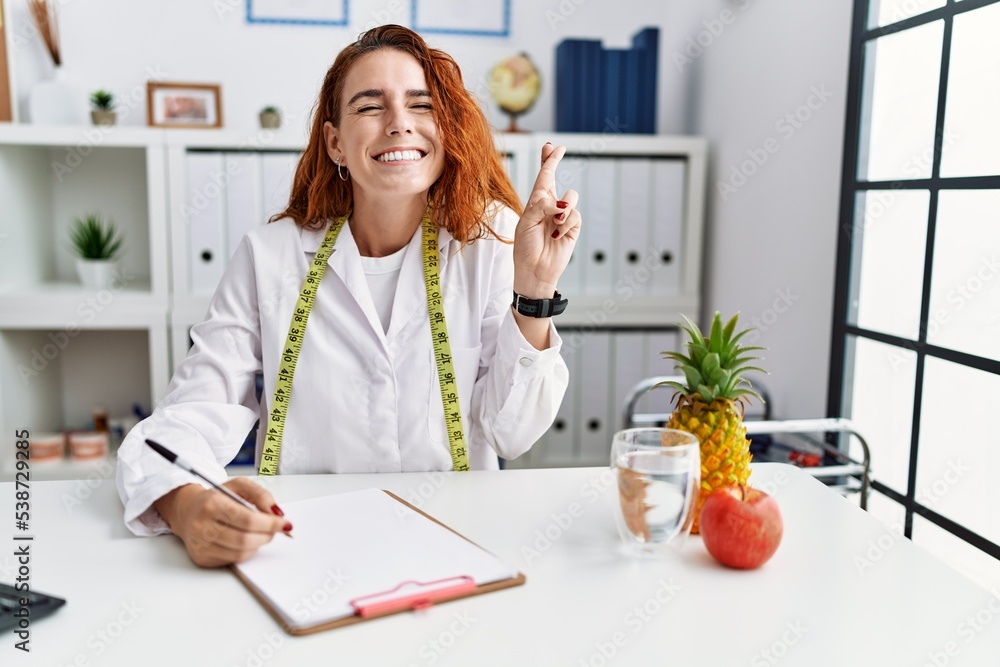 Young redhead woman nutritionist doctor at the clinic gesturing finger crossed smiling with hope and eyes closed. luck and superstitious concept.