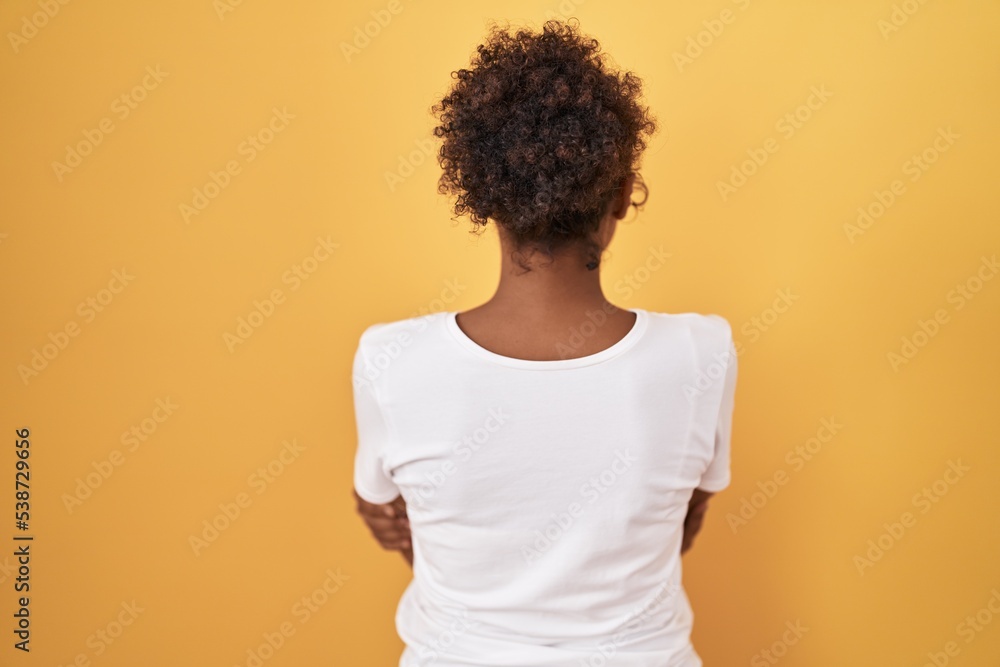 Young hispanic woman with curly hair standing over yellow background standing backwards looking away with crossed arms Young hispanic woman with curly hair standing over yellow background standing backwards looking away with crossed arms