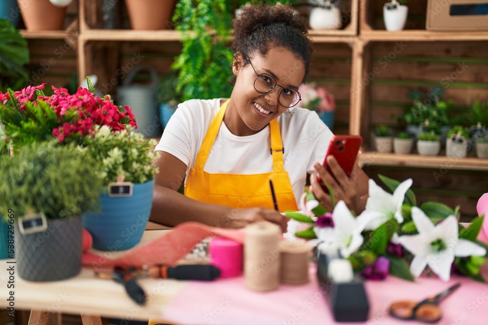 African american woman florist using smartphone writing on notebook at flower shop African american woman florist using smartphone writing on notebook at flower shop