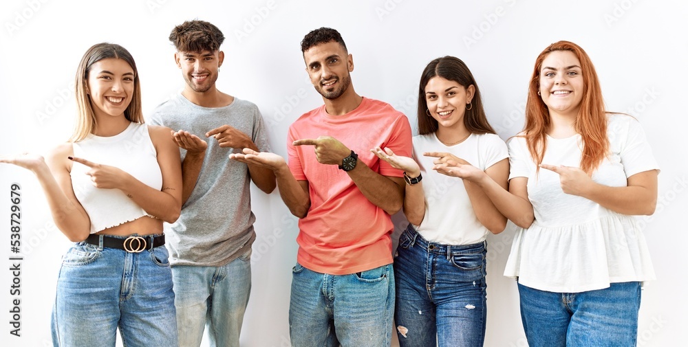Obraz premium Group of young friends standing together over isolated background amazed and smiling to the camera while presenting with hand and pointing with finger.