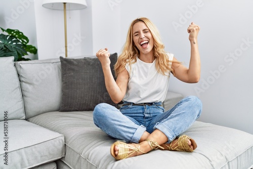 Beautiful blonde woman sitting on the sofa at home very happy and excited doing winner gesture with arms raised, smiling and screaming for success. celebration concept.